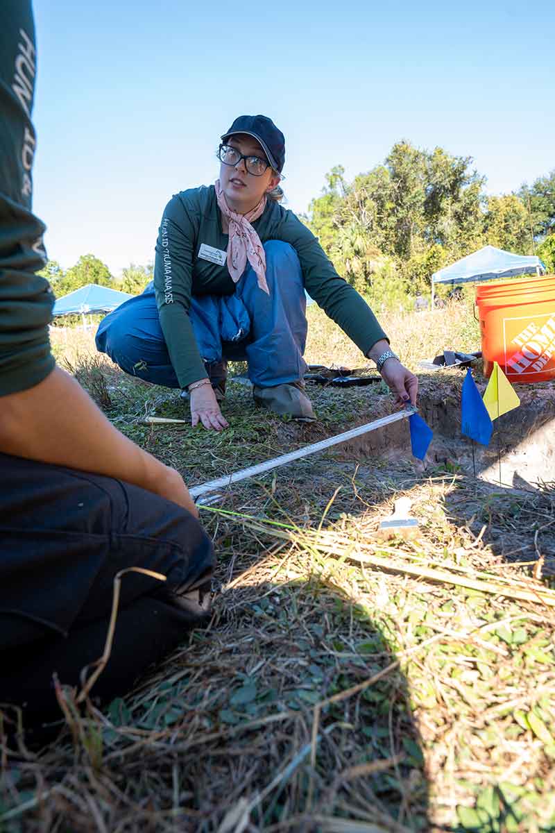A woman in glasses, a green hat and green long-sleeve shirt crouches on the ground, holding a measuring tape in one hand over a grassy area