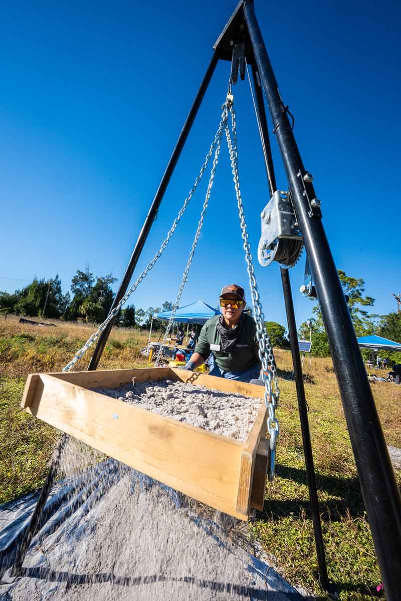 A woman in a green, long-sleeve shirt, hat and sunglasses, sifts dirt through a tripod soil sifter