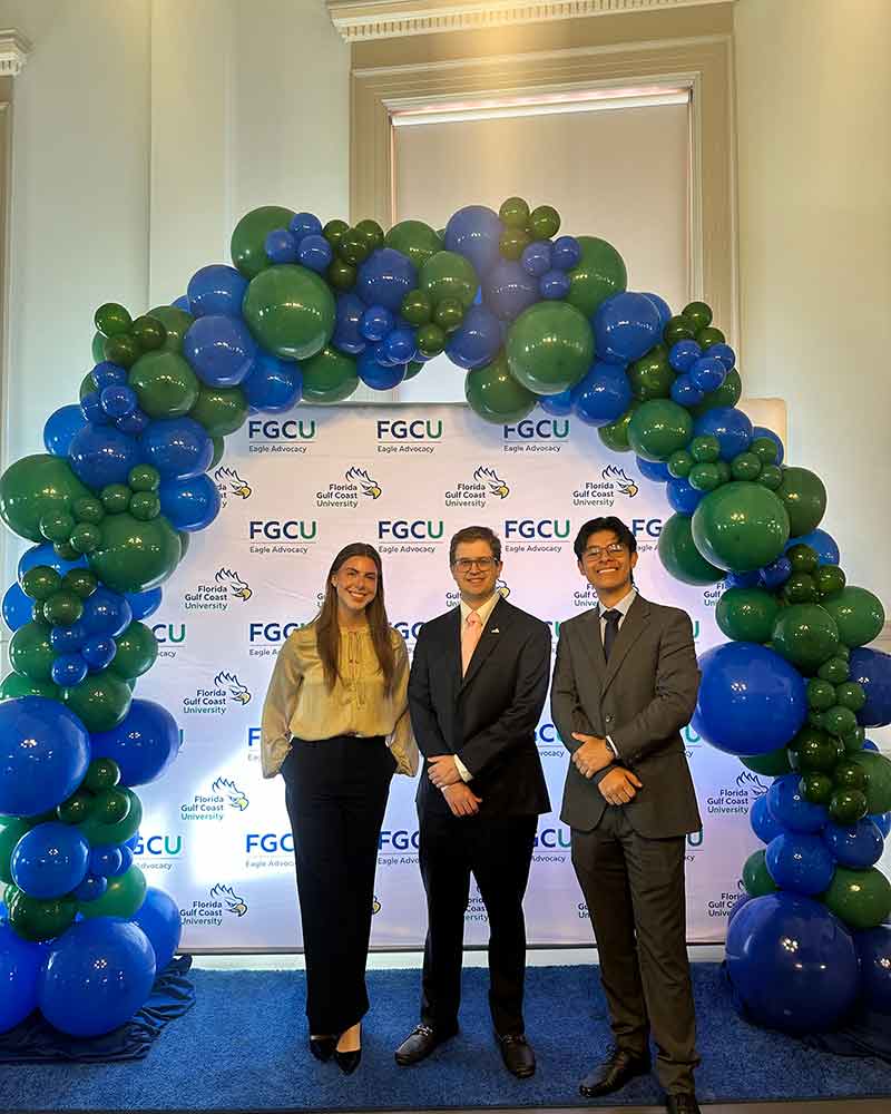 Three smiling young adults stand under a blue and green balloon arch with an FGCU step-and-repeat behind them