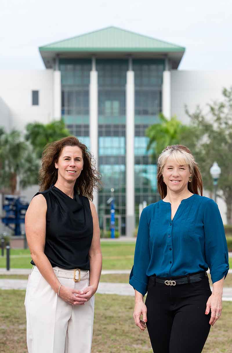 A woman with curly, dark hair, a black shirt and white pants stands with a woman with pale blonde bangs and red hair wearing a blue blouse, in front of a two-story, green-roofed building