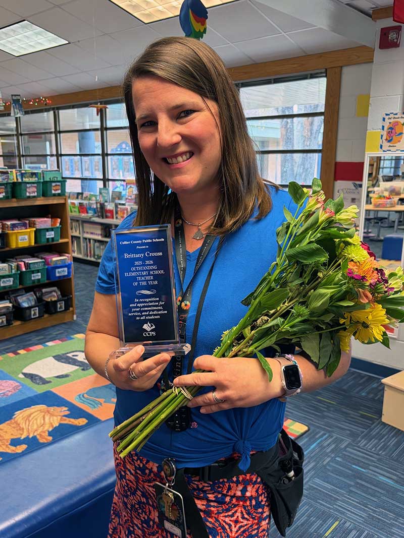 Person holding a Teacher of the Year award and a bouquet of flowers inside a classroom.