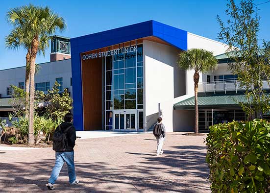 Students walk toward Cohen Student Union building with palm trees.