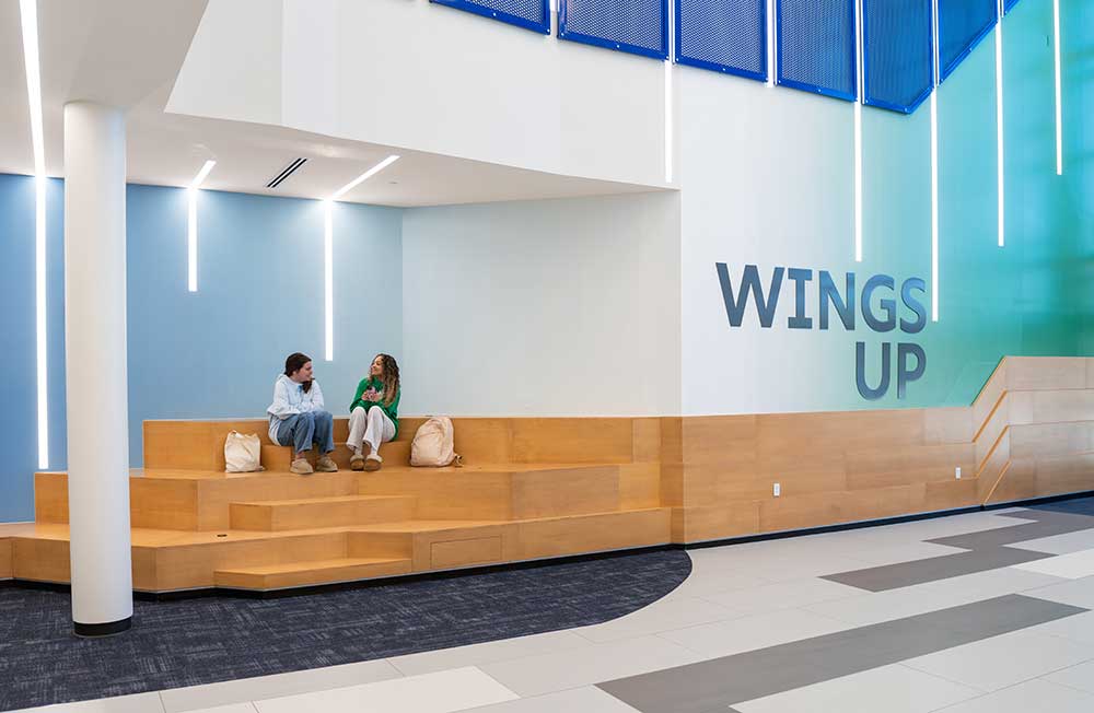Students seated on tiered wooden benches beneath a wall reading WINGS UP.