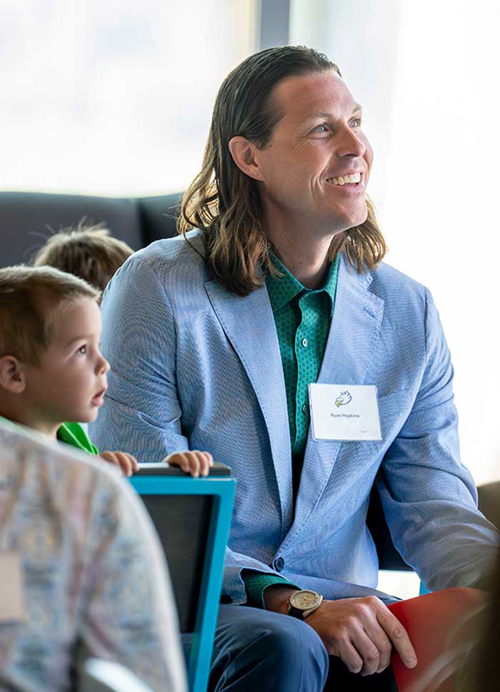 Adult in blue jacket seated at event beside a child holding a tablet.