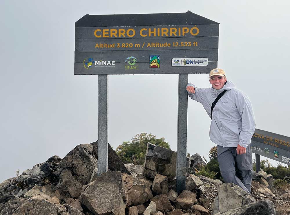 Person standing beside the Cerro Chirripó summit sign on a rocky mountaintop.