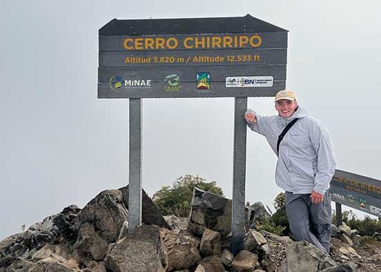 Person standing beside the Cerro Chirripó summit sign on a rocky mountaintop.