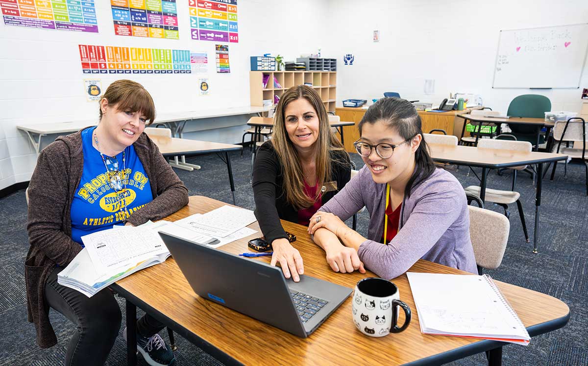 Three people collaborate at a classroom table, working on a laptop with notebooks and teaching materials.