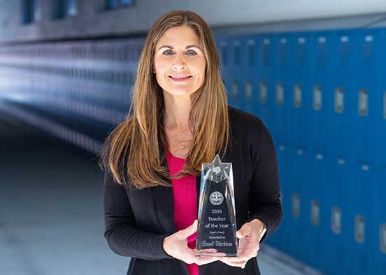 Person holding a Teacher of the Year award while standing in a hallway lined with blue lockers.
