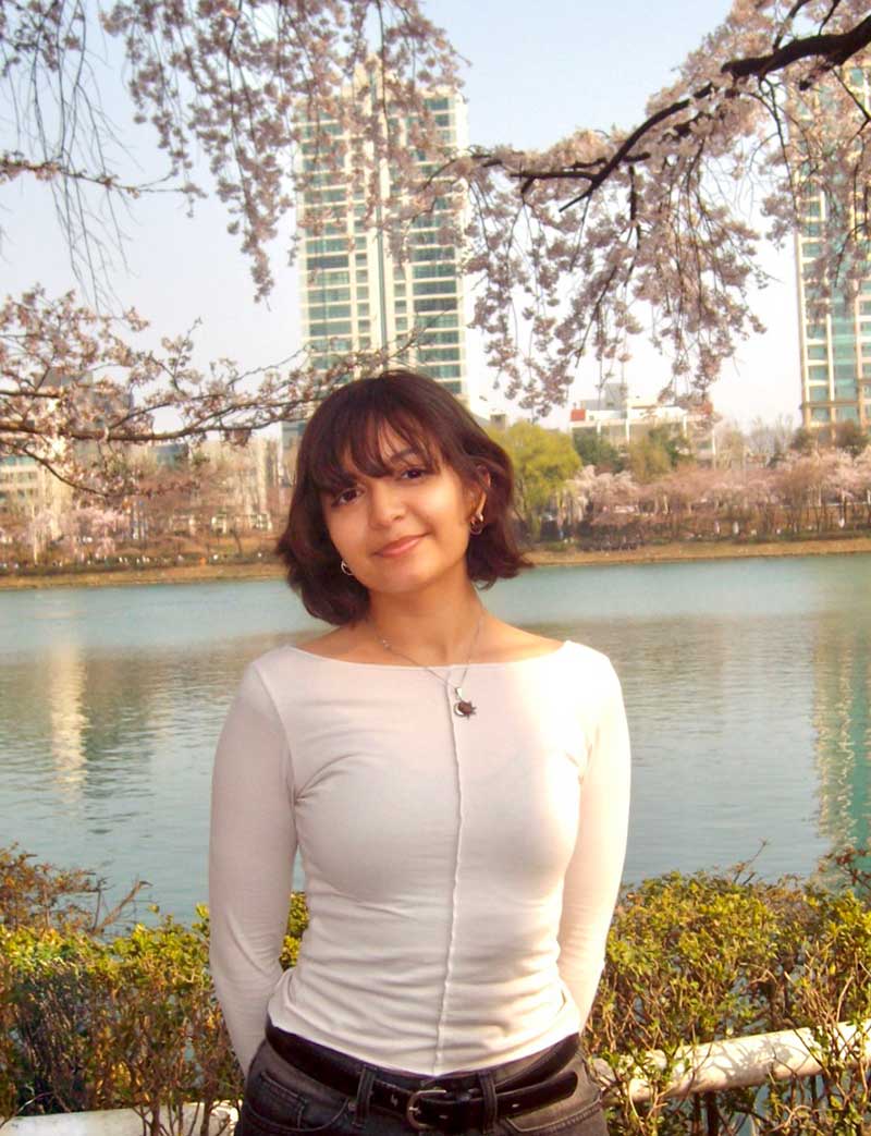 Person standing by a lakeside with cherry blossoms and tall buildings in the background.