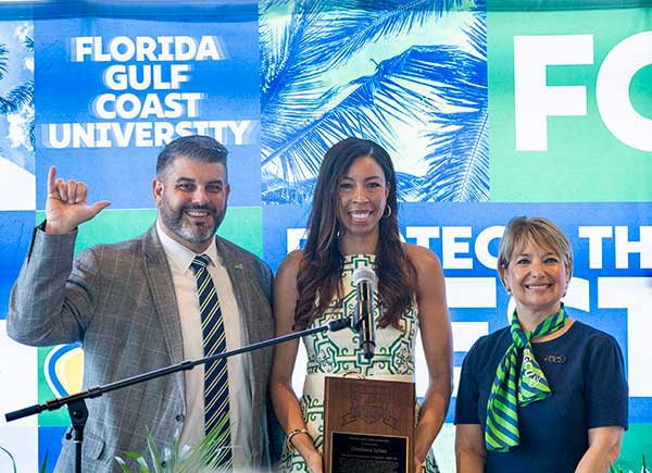 Three people pose at podium while one holds plaque during FGCU Athletics Hall of Fame ceremony.