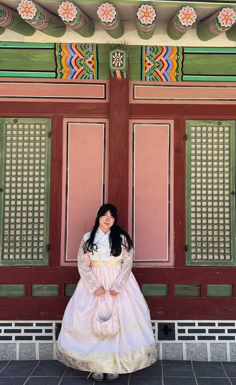 Person wearing a traditional hanbok stands before a colorful, ornate Korean building façade.
