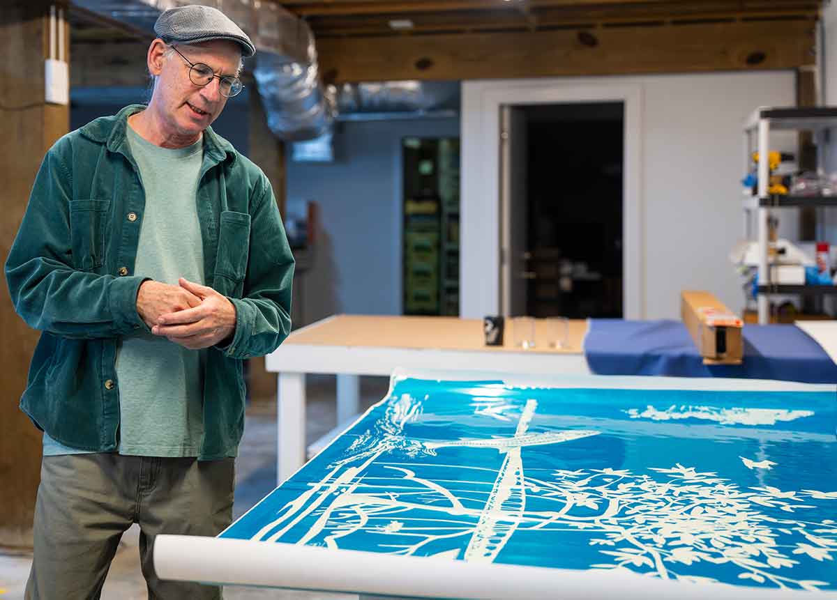 A man in a green corduroy shirt over a pale green t-shirt, in a cap and glasses, stands looking down at a large blue print featuring white, etched imagery, with tools and work surfaces visible around him