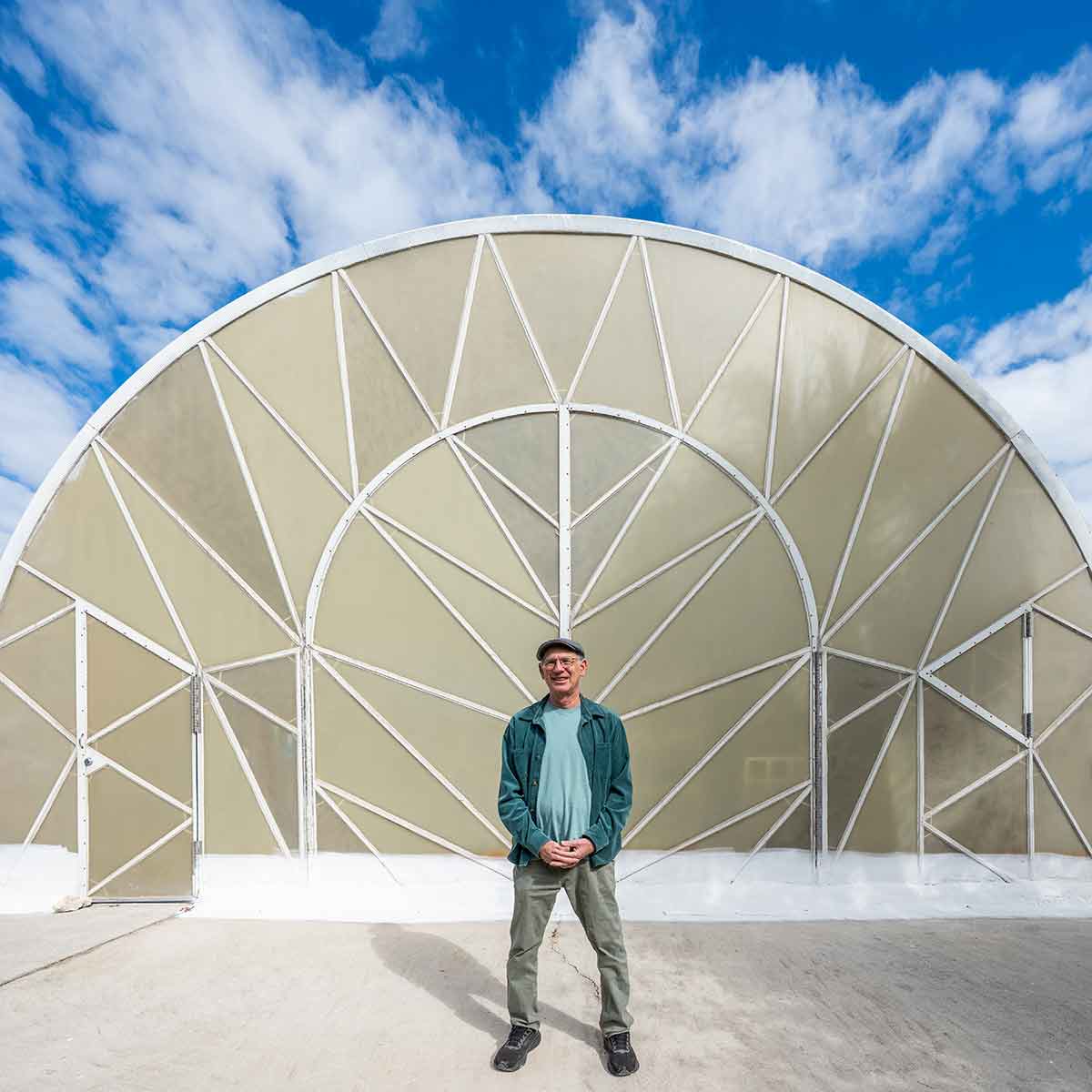 A man stands centered in front of a large, semi-circular architectural structure with white geometric framing and a pale beige surface, set against a bright blue sky with scattered clouds