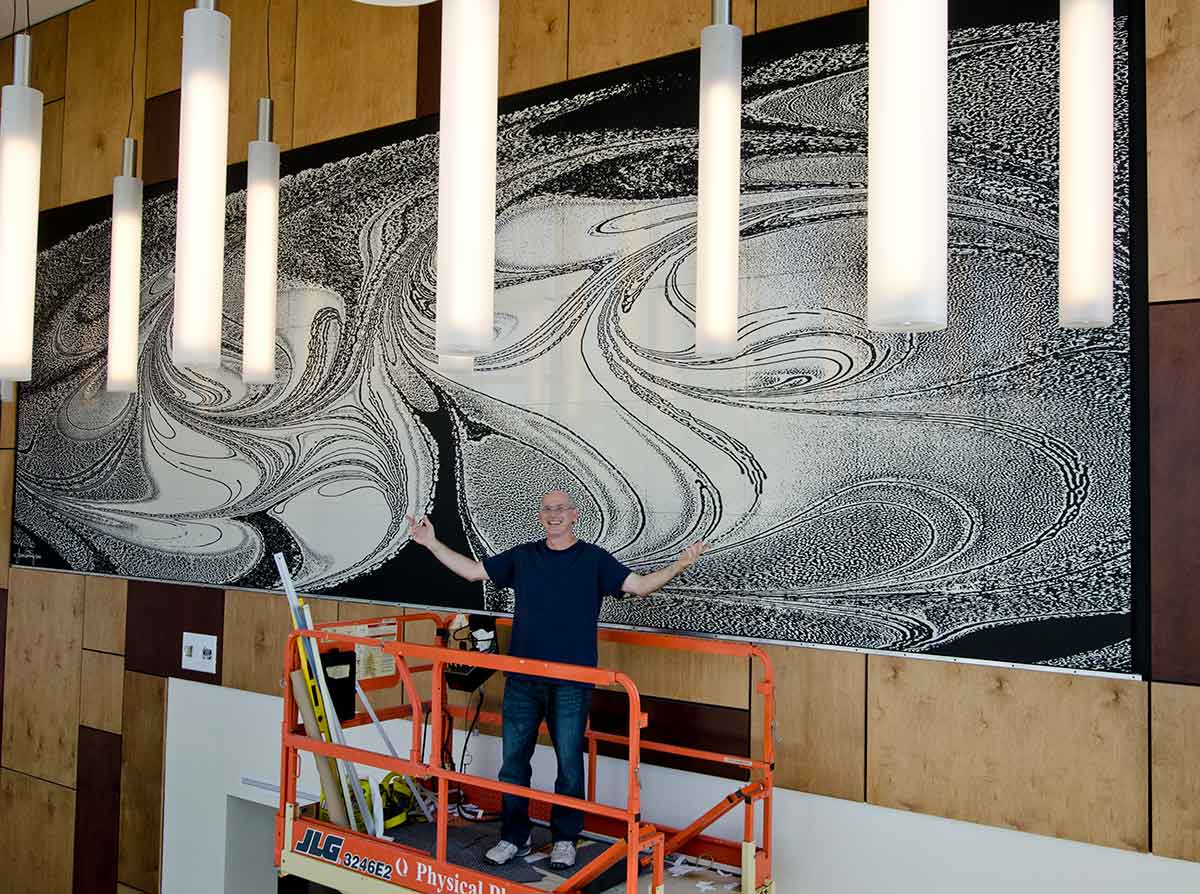 A man on an orange lift stands with his arms wide open in front of a large art installation