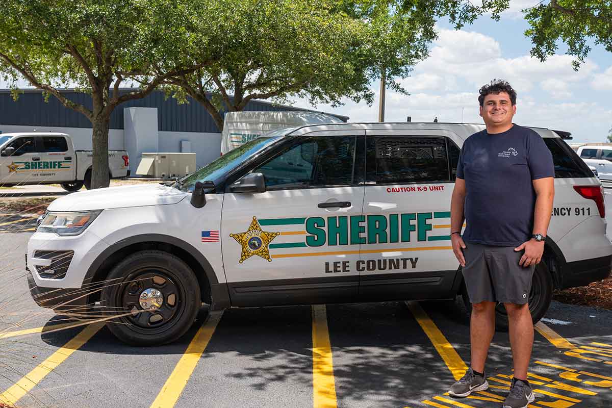 A smiling man with brown hair, a blue t-shirt with the FGCU wordmark logo and grey shorts stands next to an SUV that reads Sheriff Lee County, parked outdoors in a parking lot