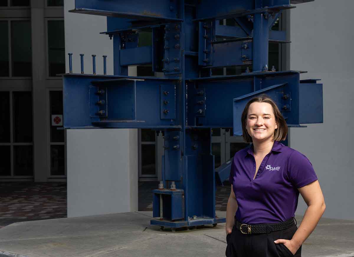 A woman in a purple, V-neck polo stands in front of the blue, metal sculpture in front of Whitaker Hall with her hands in her pockets