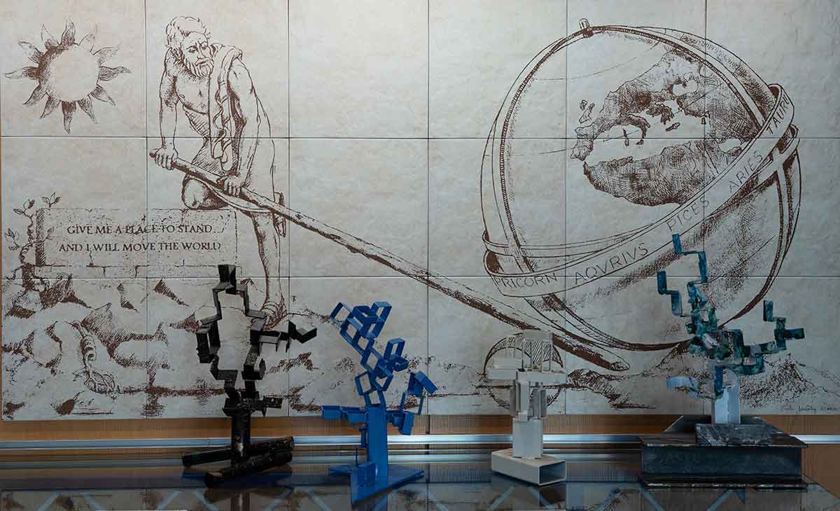 An image of a man lifting the earth with a long rod while the sun shines behind him. The image is etched on the wall behind a table with some artwork atop it