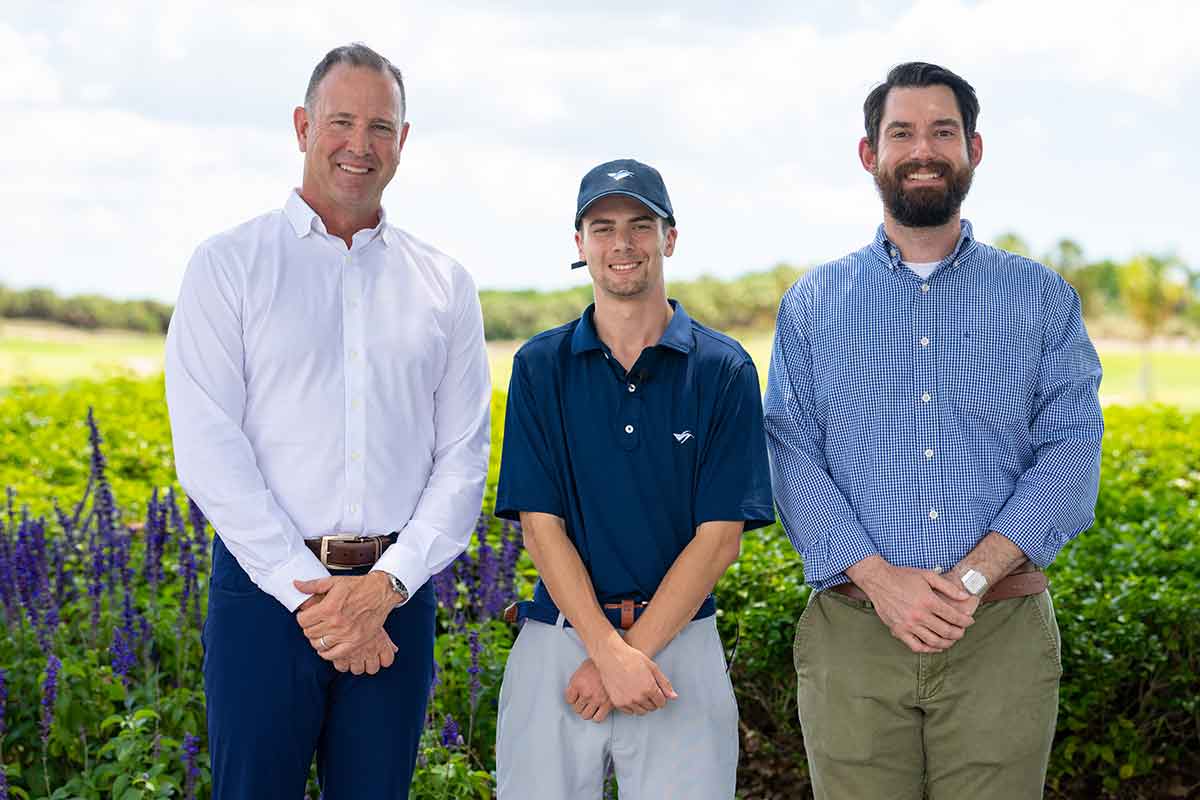 Three men stand outdoors. The older man on the far left is in a white button-down shirt and blue slacks. The younger man in the middle wears a baseball cap, navy blue polo and light-colored slacks. The man on the right has a bushy beard, light blue button-down shirt and dark tan slacks
