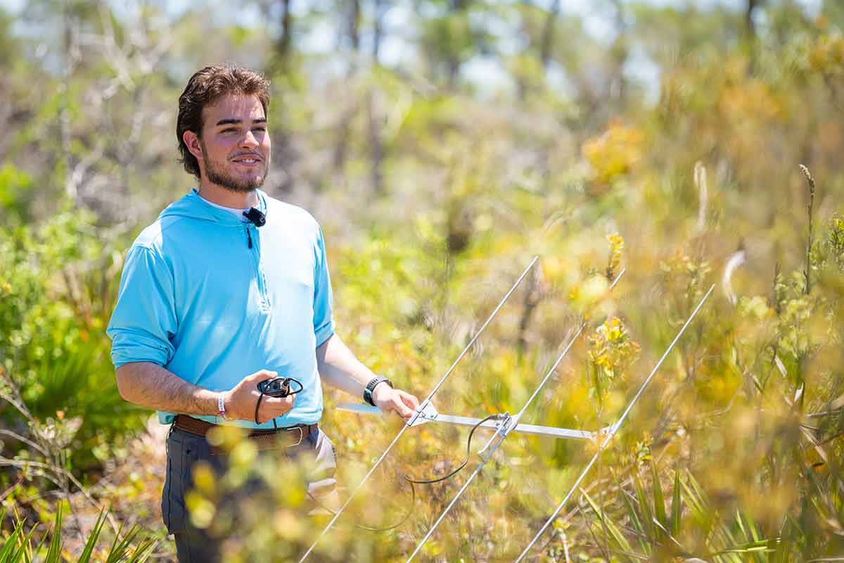 A smiling man with brown hair and a brown beard and blue, long-sleeved t-shirt stands with a metal radio telemetry device in his left hand