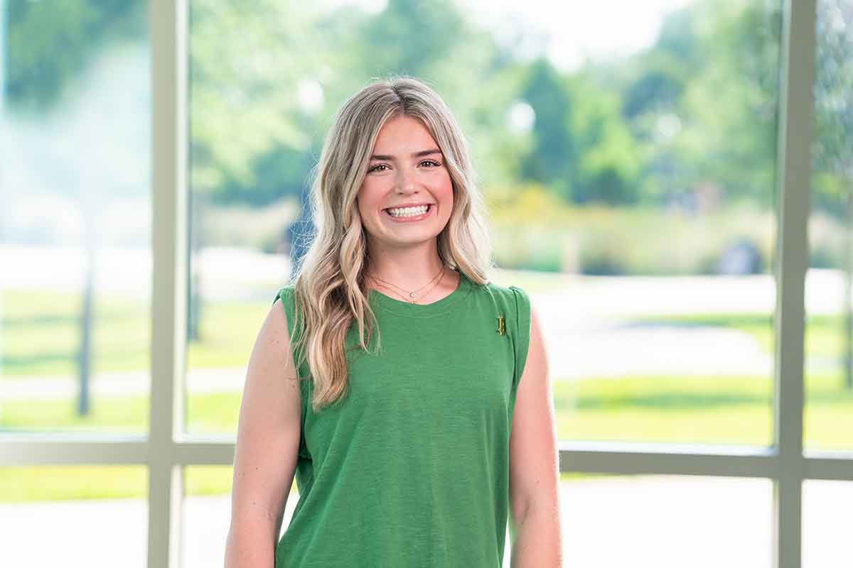 A smiling, blonde woman in a sleeveless, green top, stands in front of a large window on a sunny day