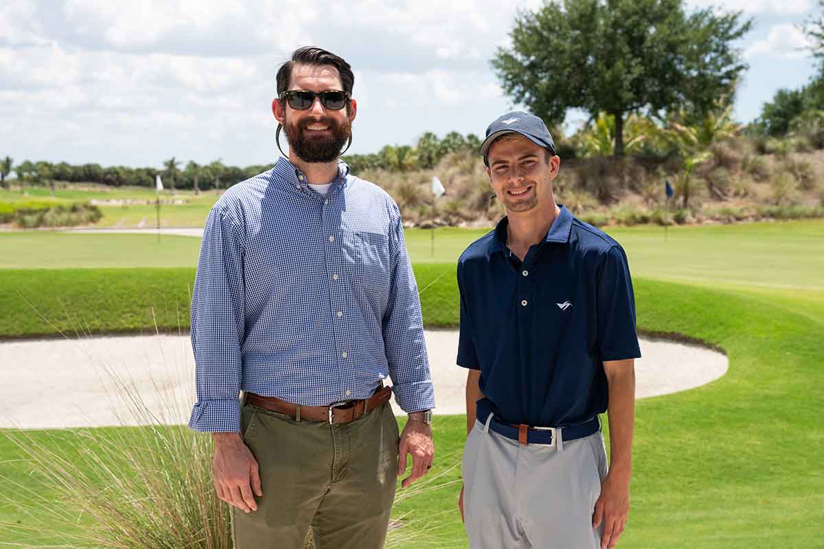Two men stand outdoors in front of a golf course sand trap. The man on the left is a head taller, with a bushy beard, sunglasses. The younger man on the right wears a baseball cap, navy blue polo and light-colored slacks