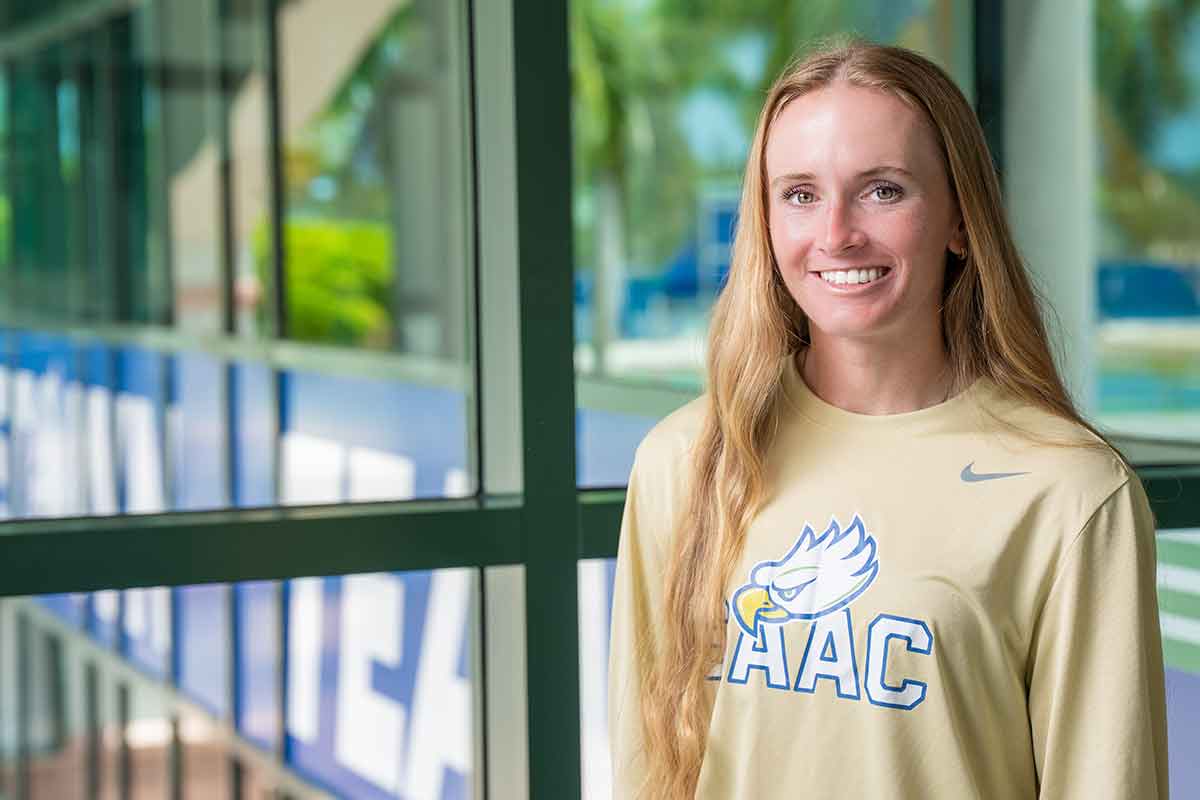 A smiling, blonde woman with a light yellow, long-sleeved t-shirt that reads SAAC and has the FGCU Athletics eagle head logo stands in front of a window