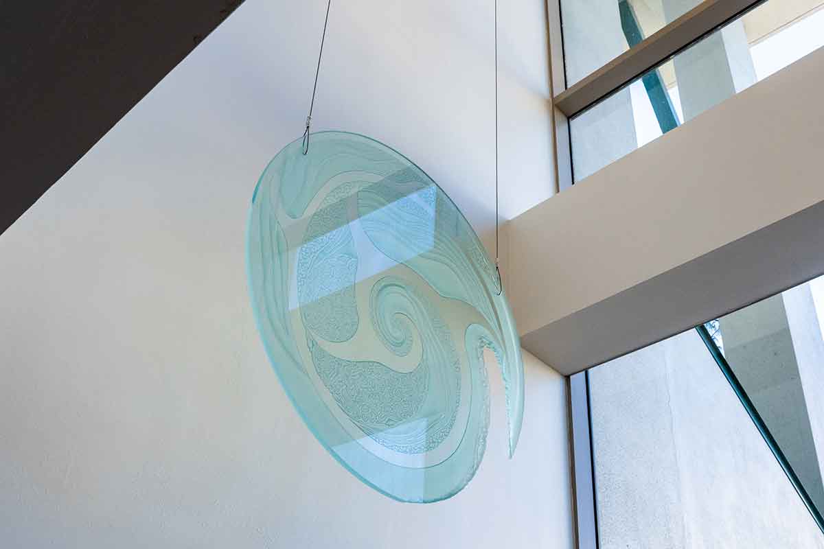 A photo of a large, round, flat piece of glass with etchings of swirling designs, hanging from the ceiling