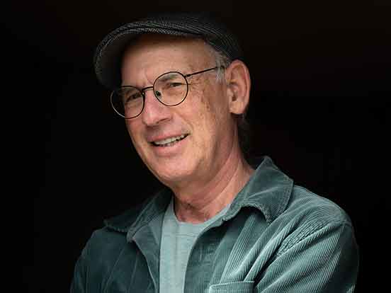 A man in a green corduroy shirt over a pale green t-shirt, in a cap and glasses, stands against a black background