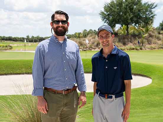 Two men stand outdoors in front of a golf course sand trap. The man on the left is a head taller, with a bushy beard, sunglasses. The younger man on the right wears a baseball cap, navy blue polo and light-colored slacks