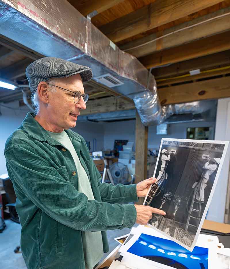 A man in a green corduroy shirt over a pale green t-shirt, in a cap and eyeglasses, stands in a basement workroom, holding a black and white photo of a man and his own image reflected in a granite wall