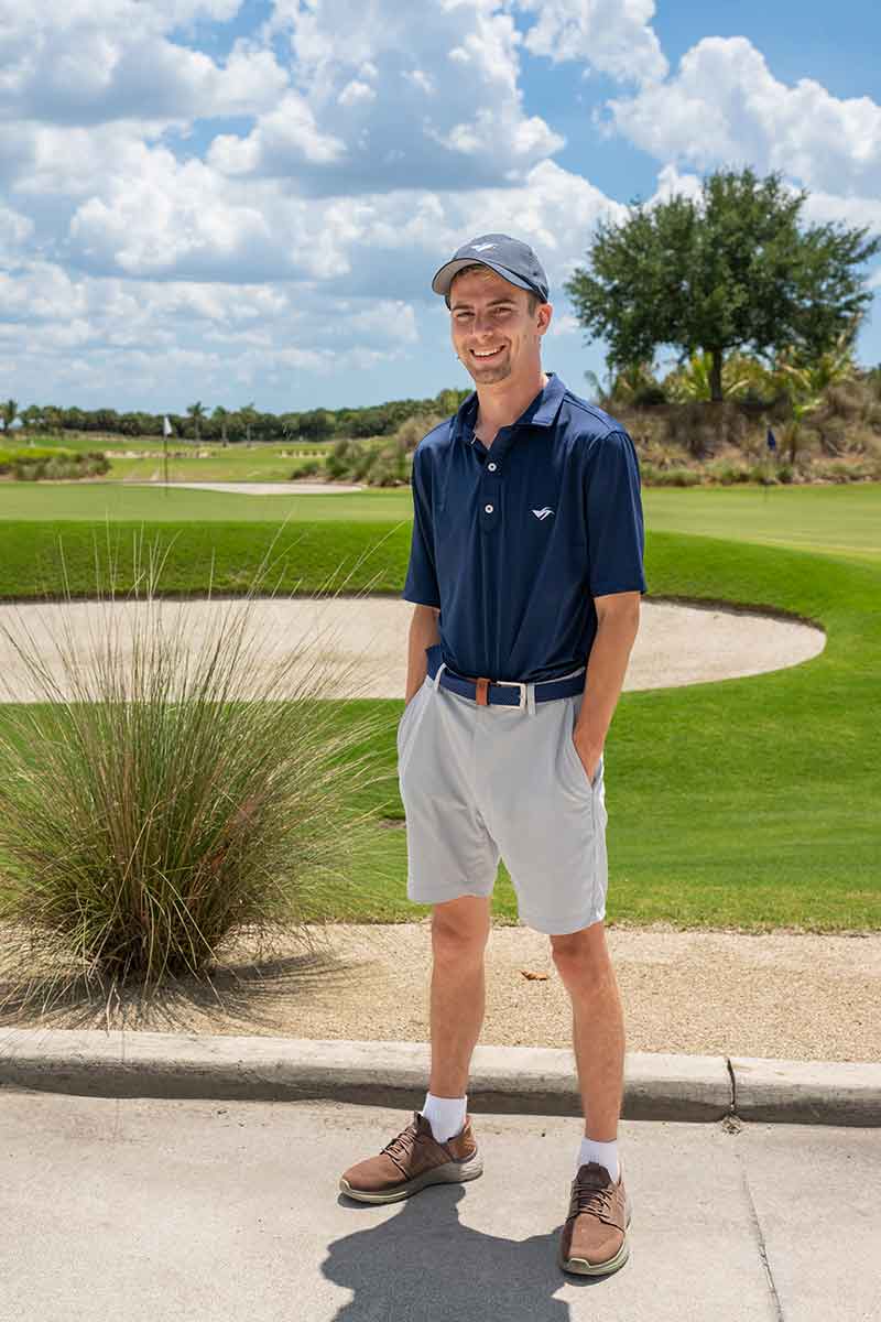 Standing outdoors in front of a golf course sand trap, a young man wears a baseball cap, navy blue polo and light-colored slacks