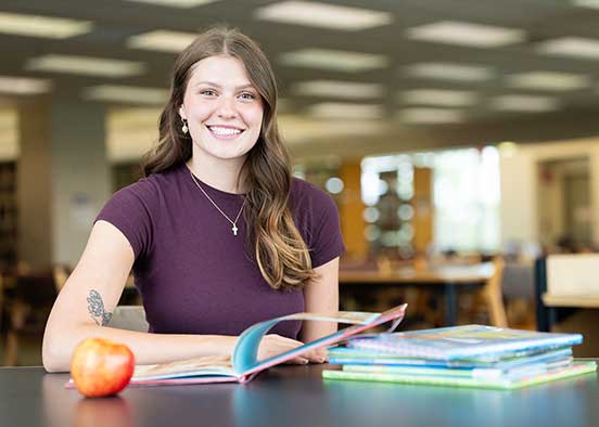 FGCU education student with textbooks and apple in campus library