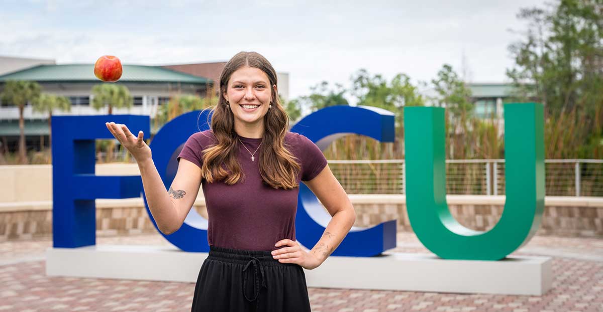 FGCU education student holding apple in front of Florida Gulf Coast University sign