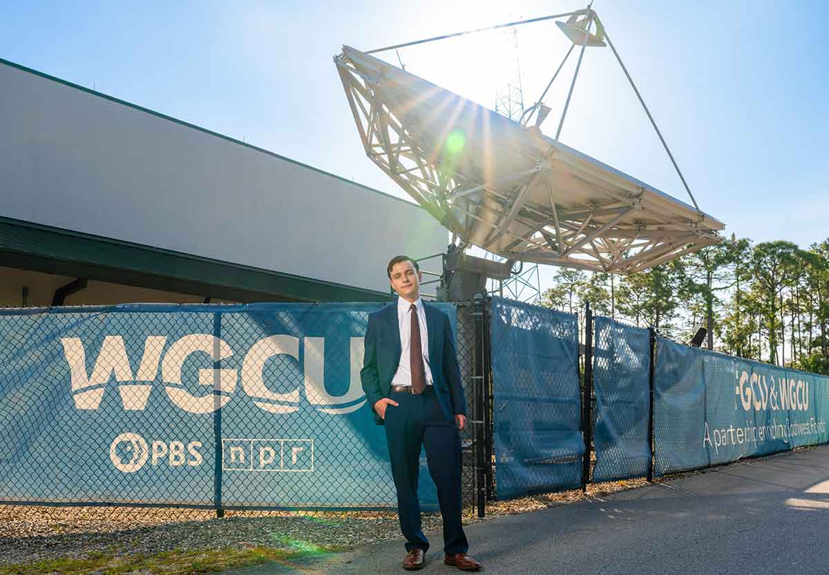 A young man in a suit and tie stands with one hand in his pocket in front of a WGCU-branded fence and the WGCU satellite dish