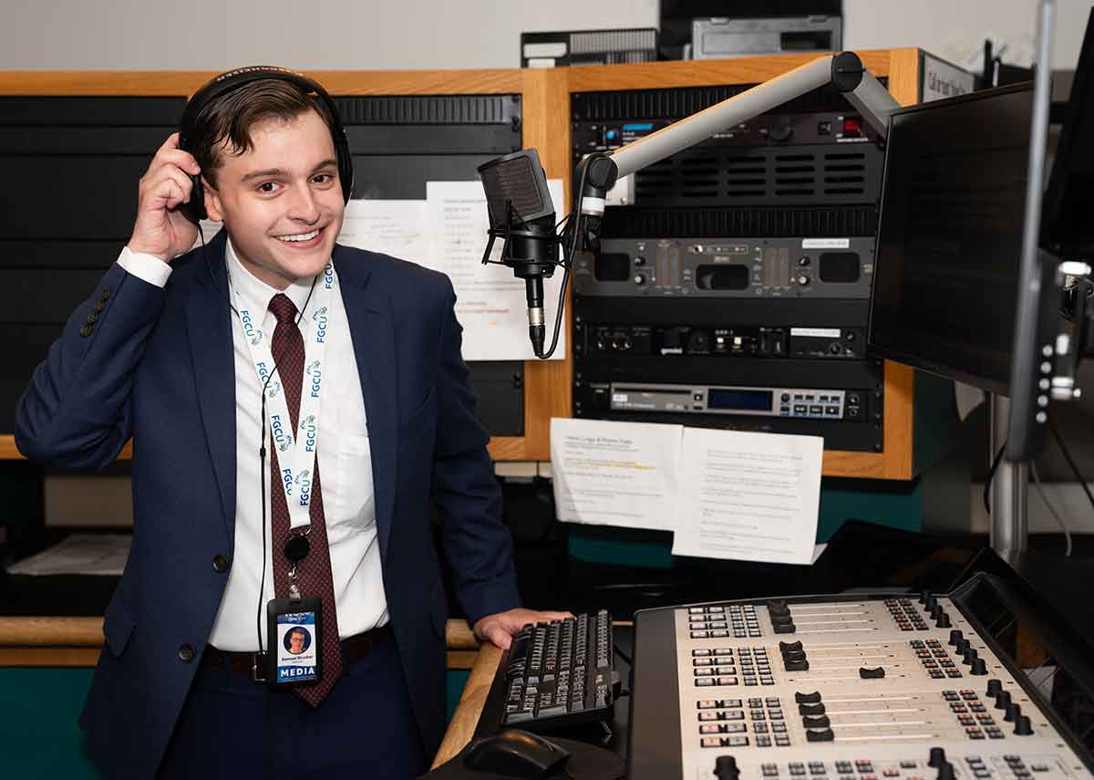 A young man in a suit and tie removes the right side of his headphones and smiles at the camera from behind the desk of a radio station