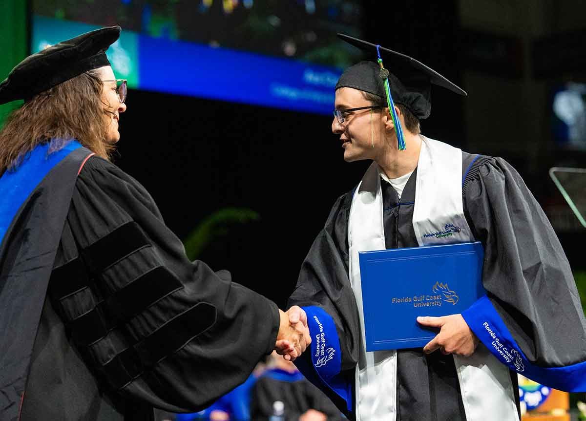 A young man in a cap and gown holds his FGCU diploma in his left hand and shakes the hand of a woman in a cap and gown at graduation