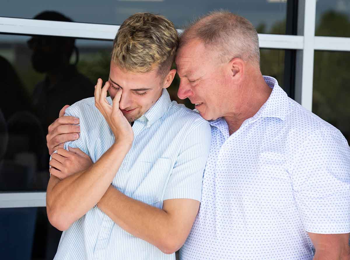 A young man in a white shirt wipes away tears as an older man side hugs him