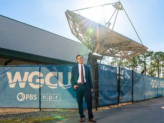 A young man in a suit and tie stands with one hand in his pocket in front of a WGCU-branded fence and the WGCU satellite dish