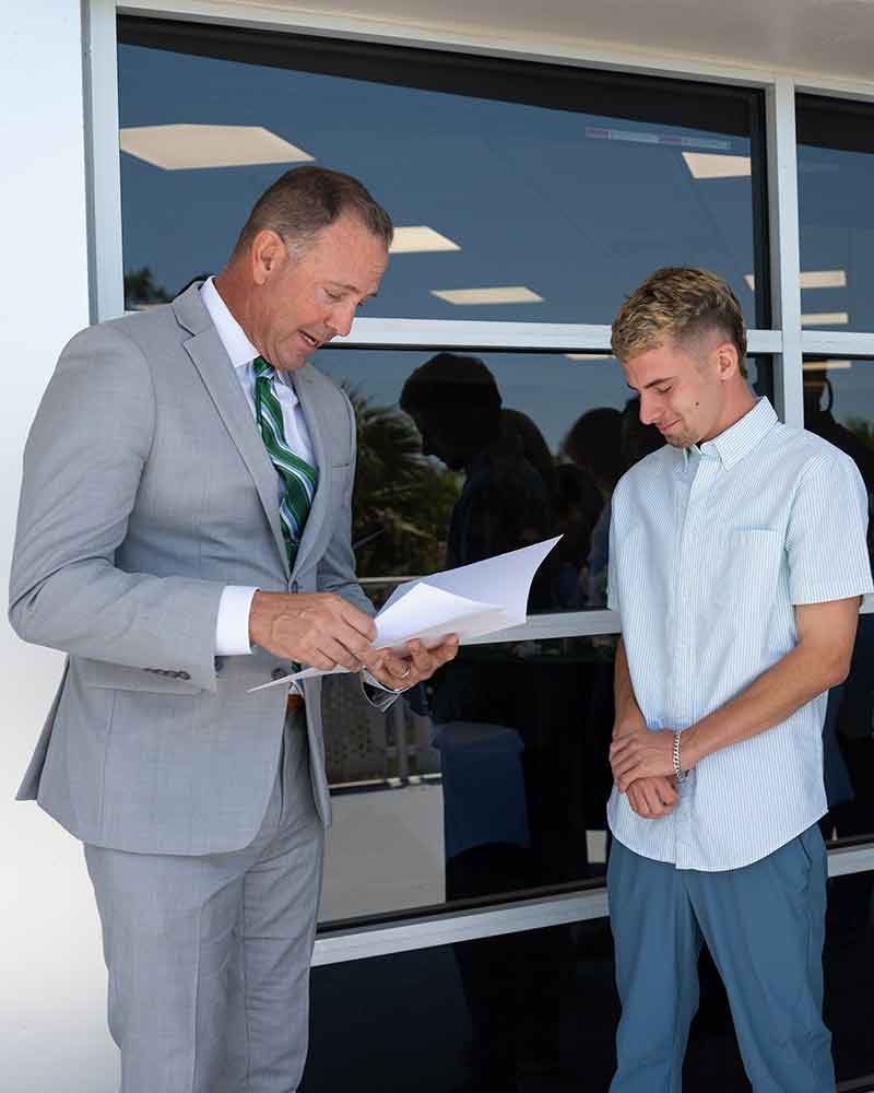 A man in a grey suit reads from a piece of paper while a young man in a white shirt and blue pants stands before him