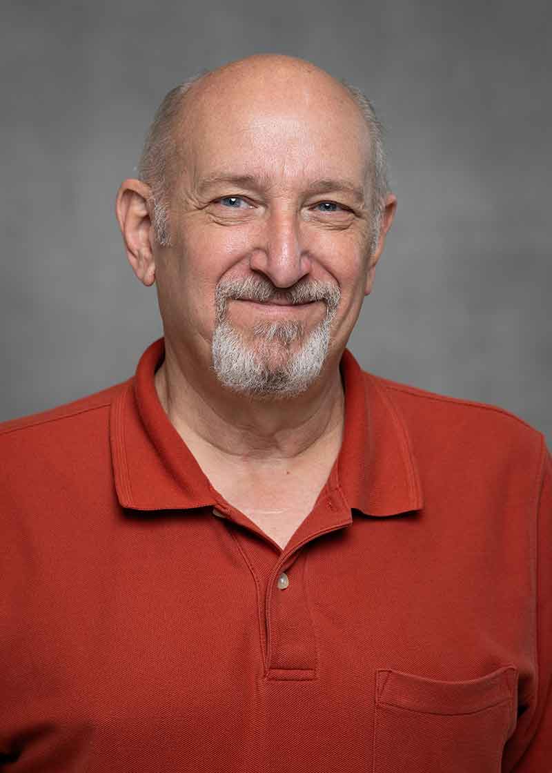 A headshot of an older man with a slat-and-pepper goatee in an orange polo shirt