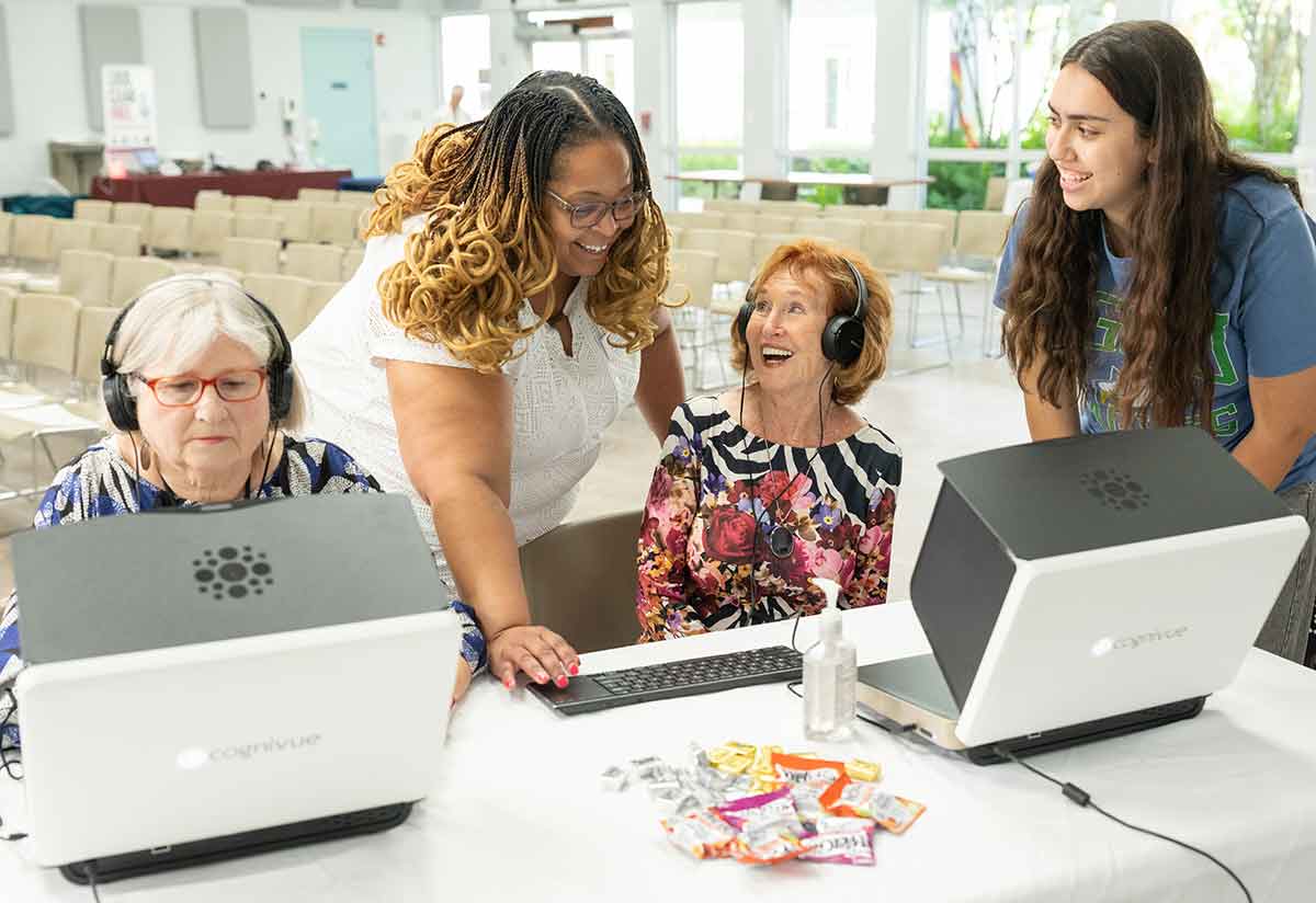 Two older women are seated at computer screens while a professor in a white dress and student in a blue FGCU shirt look on