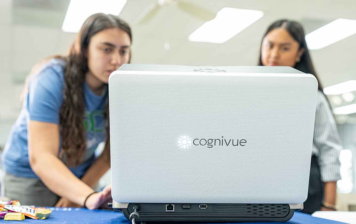 Two young women, one in a blue shirt and one in a white button-down shirt, stand looking at a computer with Cognivue written on the front