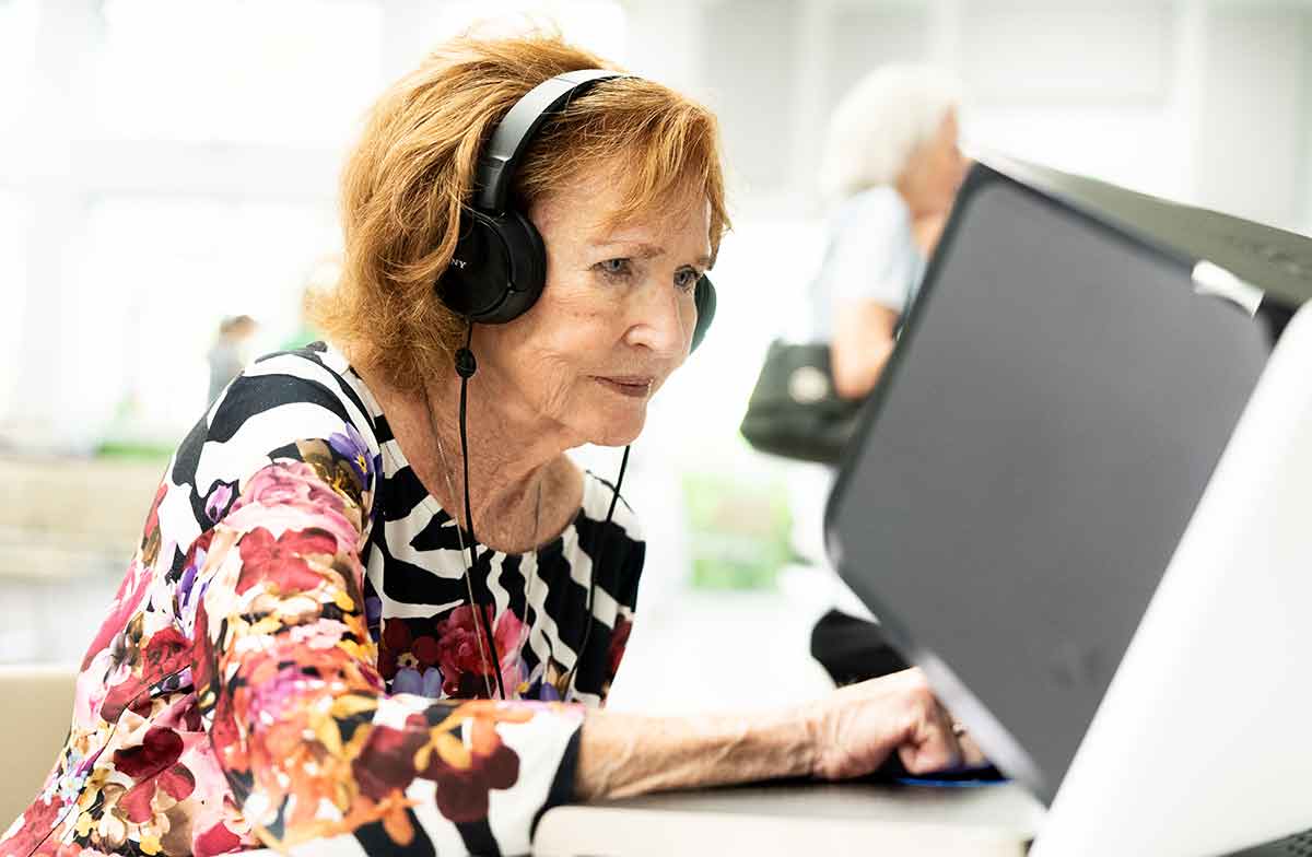 A red-headed older woman in a black, pink and white shirt sits in front of a computer