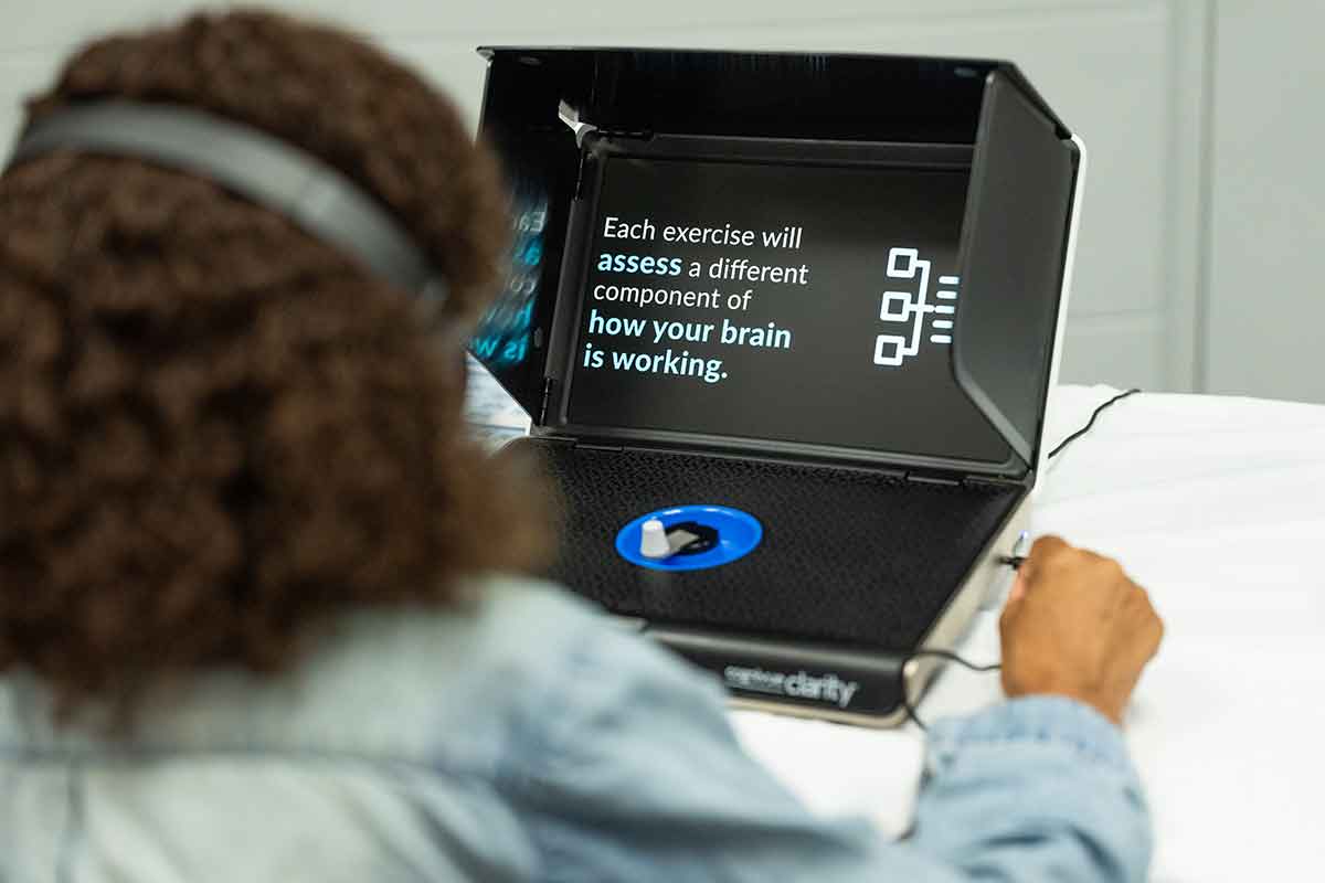 A woman with curly brown hair has her back to the camera as she works at a computer. The screen reads, Each exercise will assess a different component of how your brain is working