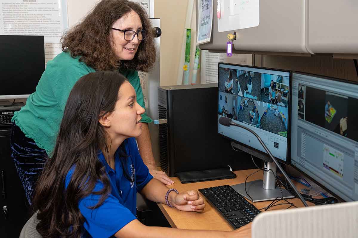 A brunette woman wearing glasses and a green top stands behind a seated young woman in a blue top as they both watch computer monitors with CCTV footage of a mother and child
