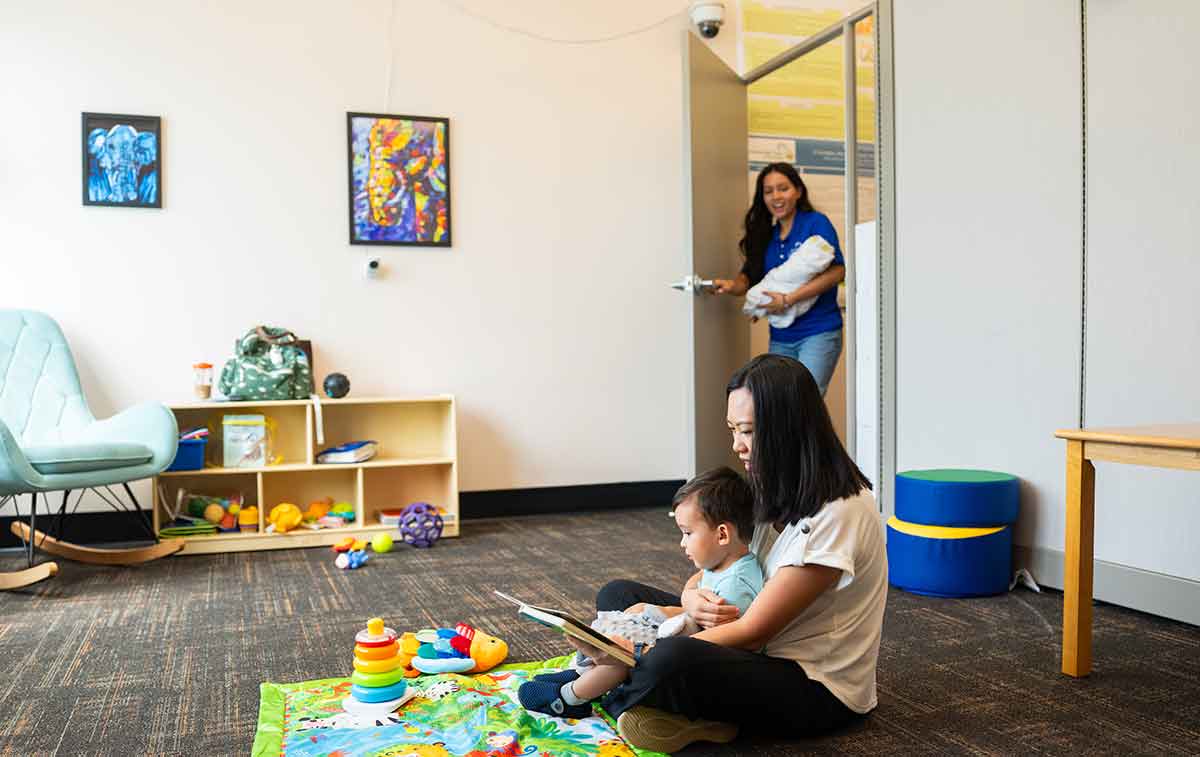 A mom and toddler sit on the floor playing as a young woman in a blue shirt enters through an open door with a baby doll wrapped in a blanket in her arms