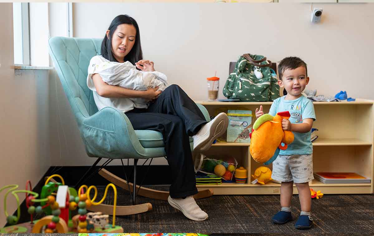 A woman in a white shirt and black pants sits in a green upholstered rocking chair, holding a lifelike baby doll in her arms, as her toddler looks on