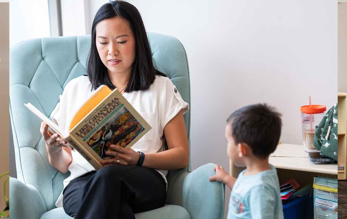 A woman in a white shirt and black pants sits in a green upholstered rocking chair, reading a book, as her toddler looks on