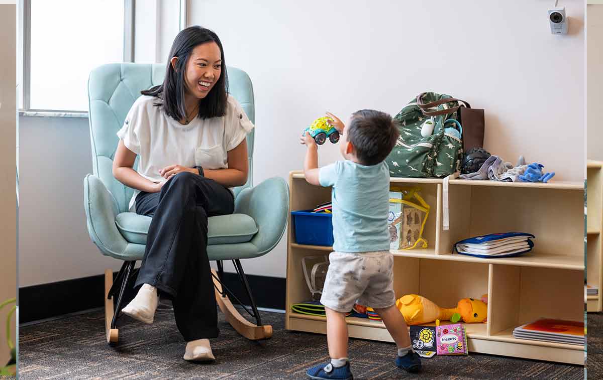 A woman in a white top and jeans sits in a green upholstered rocking chair, smiling down at her toddler playing with toys on a low bookcase