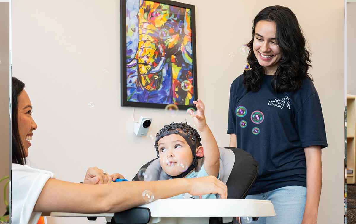 A mom in profile on the left, with a baby in a high chair and skull cap centered, and a young woman in a blue t-shirt standing behind him